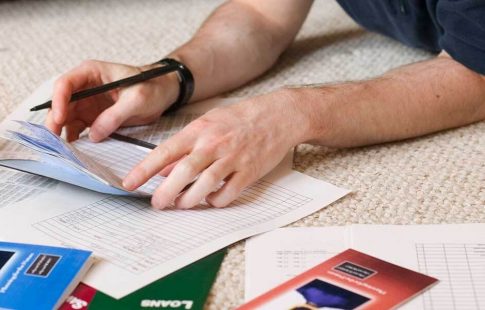 Man's hands holding a checkbook near a student loan pamphlet, scattered papers, and a calculator