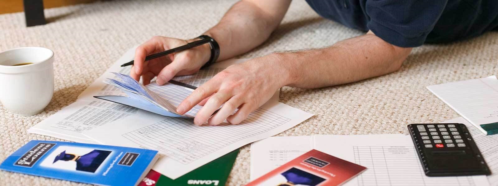 Man's hands holding a checkbook near a student loan pamphlet, scattered papers, and a calculator