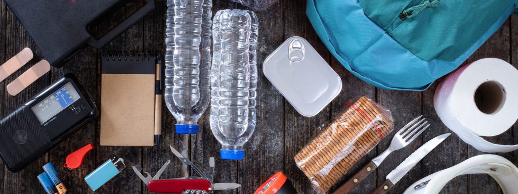 A group of home emergency survival kit supplies including water bottles, canned food, a radio, band-aids, and batteries