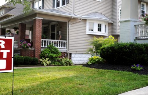 "For rent" sign in a front lawn next to a row of historic homes with porches.