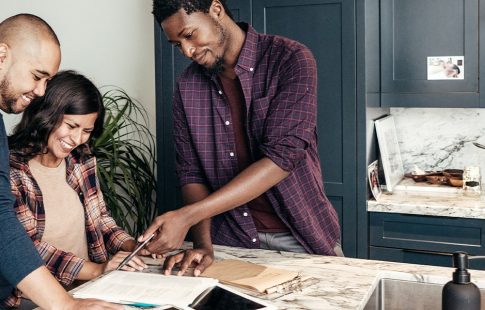 A couple looks at paperwork in a kitchen with a real estate agent. Finding an agent who works in the area where you plan to buy a home is an item that can be added to a buying a home checklist.