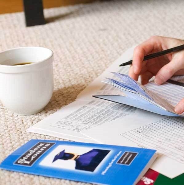 Man's hands holding a checkbook near a student loan pamphlet, scattered papers, and a calculator