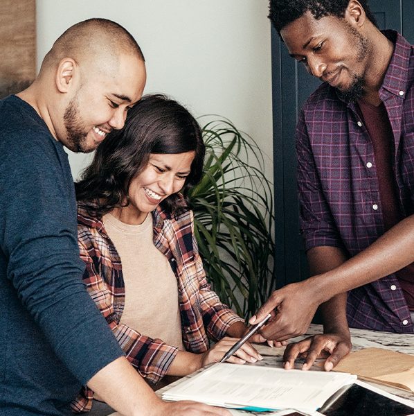 A couple looks at paperwork in a kitchen with a real estate agent. Finding an agent who works in the area where you plan to buy a home is an item that can be added to a buying a home checklist.