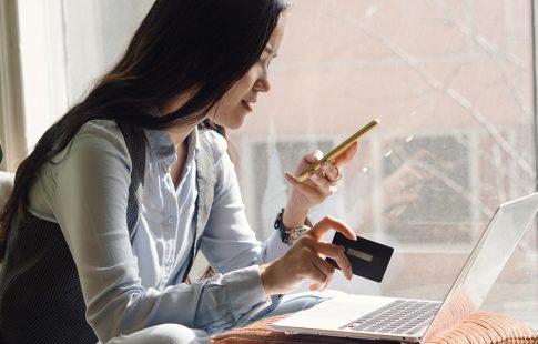 Woman holding credit card and smart phone in front of laptop, wondering, "can you consolidate credit card debt into your mortgage?"