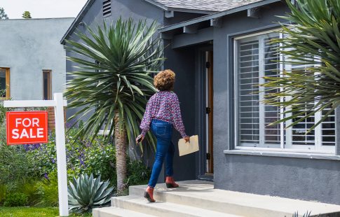Woman walking past a "for sale" sign and into a house. She has a folder in her hand to help her through the home buying process.