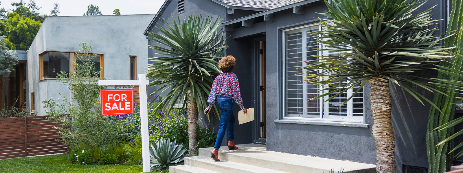 Woman walking past a "for sale" sign and into a house. She has a folder in her hand to help her through the home buying process.
