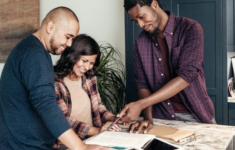 A couple looks at paperwork in a kitchen with a real estate agent. Finding an agent who works in the area where you plan to buy a home is an item that can be added to a buying a home checklist.