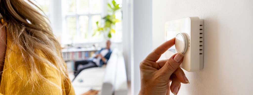 A woman adjusts a thermostat to a temperature recommended by experts on summer energy saving tips while a blurred man sits on a couch in the background