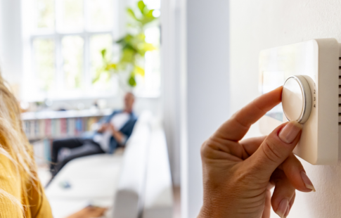 A woman adjusts a thermostat to a temperature recommended by experts on summer energy saving tips while a blurred man sits on a couch in the background