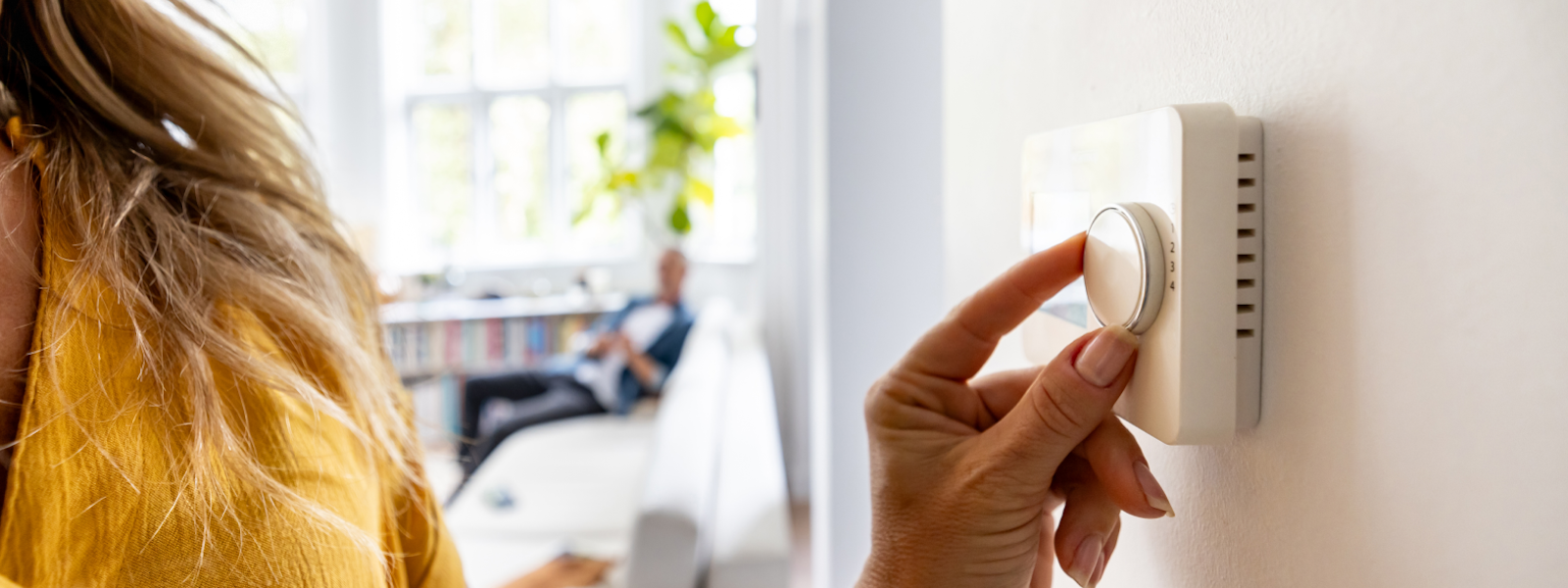 A woman adjusts a thermostat to a temperature recommended by experts on summer energy saving tips while a blurred man sits on a couch in the background